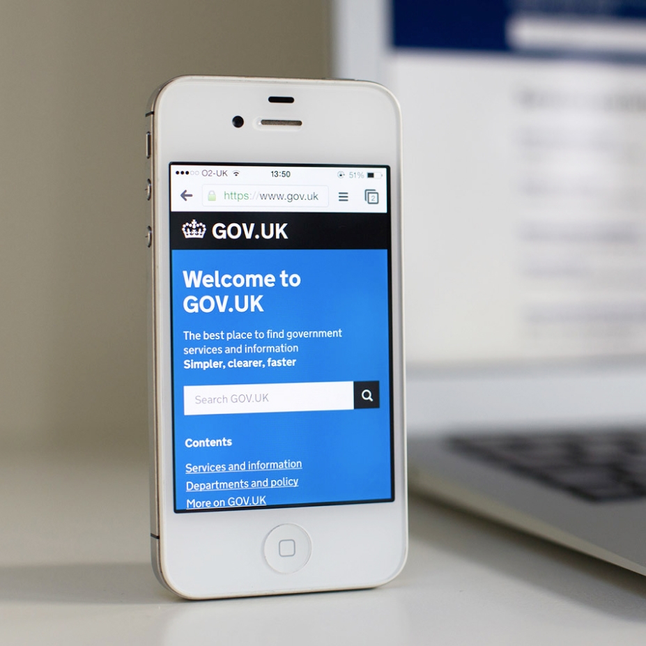 A photograph of a white iPhone 5S showing the GOV.UK homepage on its screen. An out of focus laptop can be seen in the background