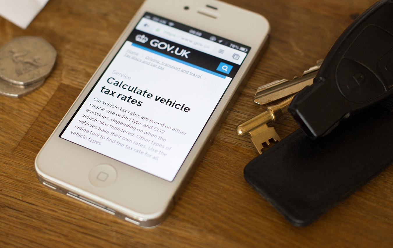 A photograph of an iPhone 5S showing the GOV.UK 'Calculate vehicle tax rates' page on its screen. The phone is next to some car keys and coins.